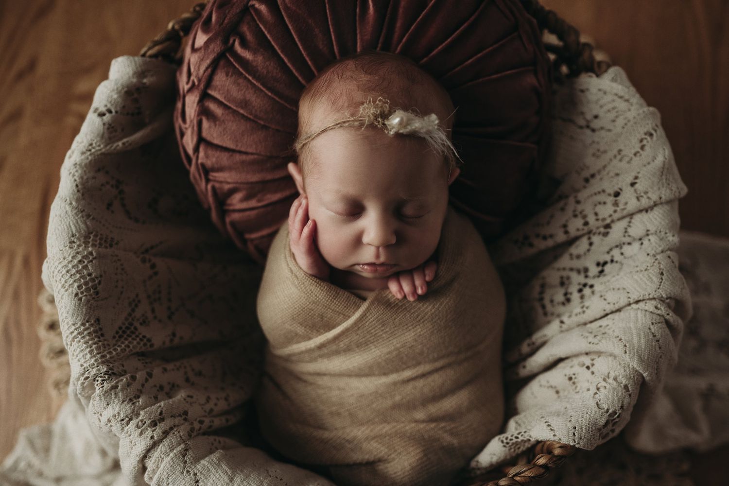 Sleeping infant wrapped in beige fabric lying in brown circular cushion on wooden floor.