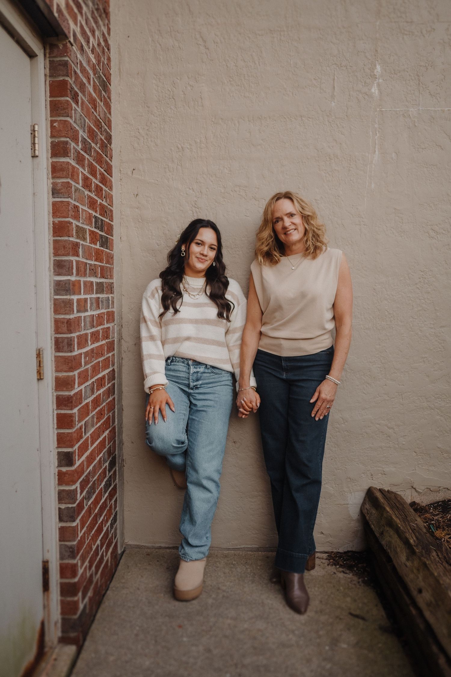Two people posing together against a beige stucco wall and brick column while wearing casual attire.