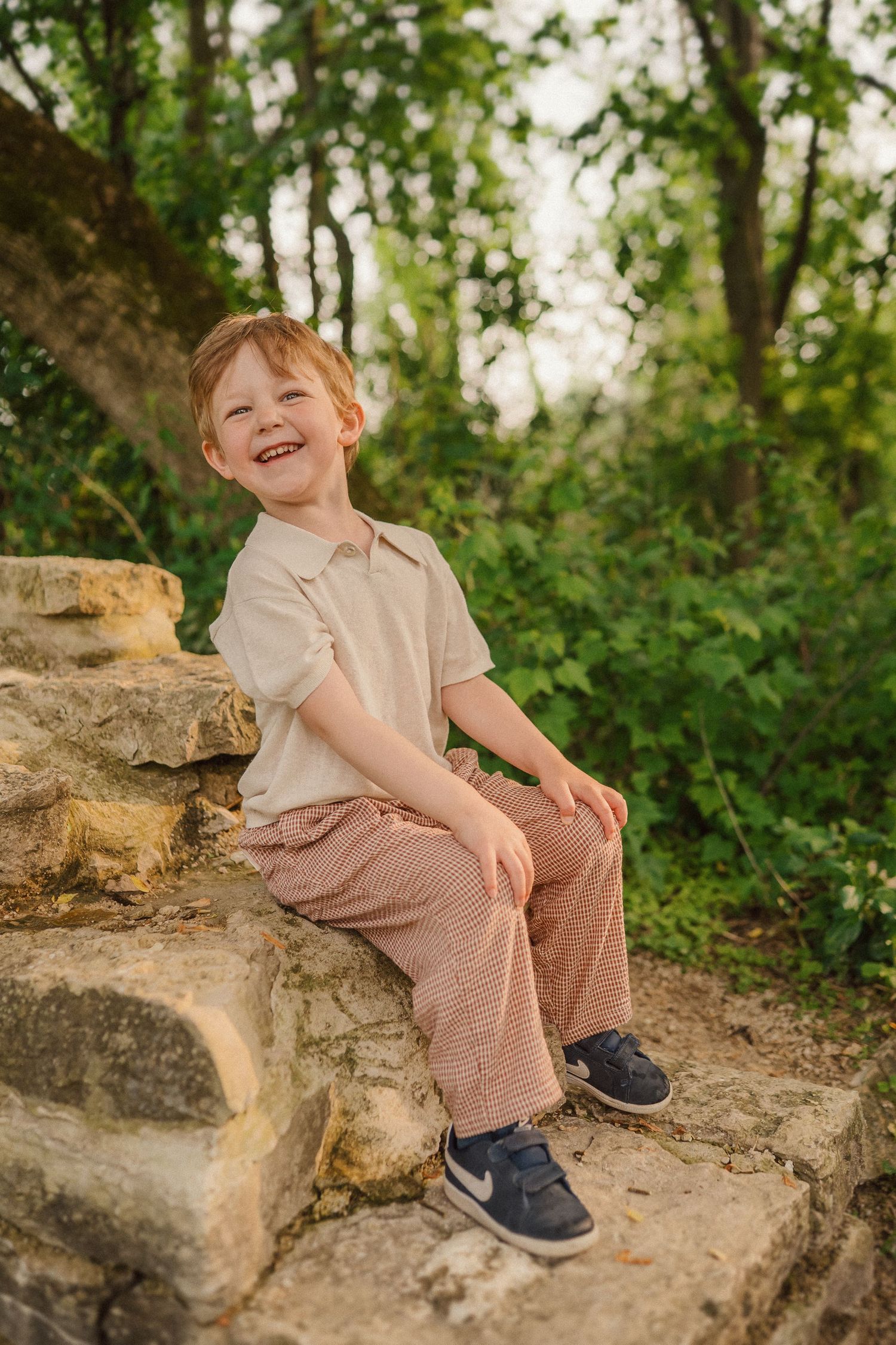 A child in beige clothing sits on stone steps outdoors against a lush green background.