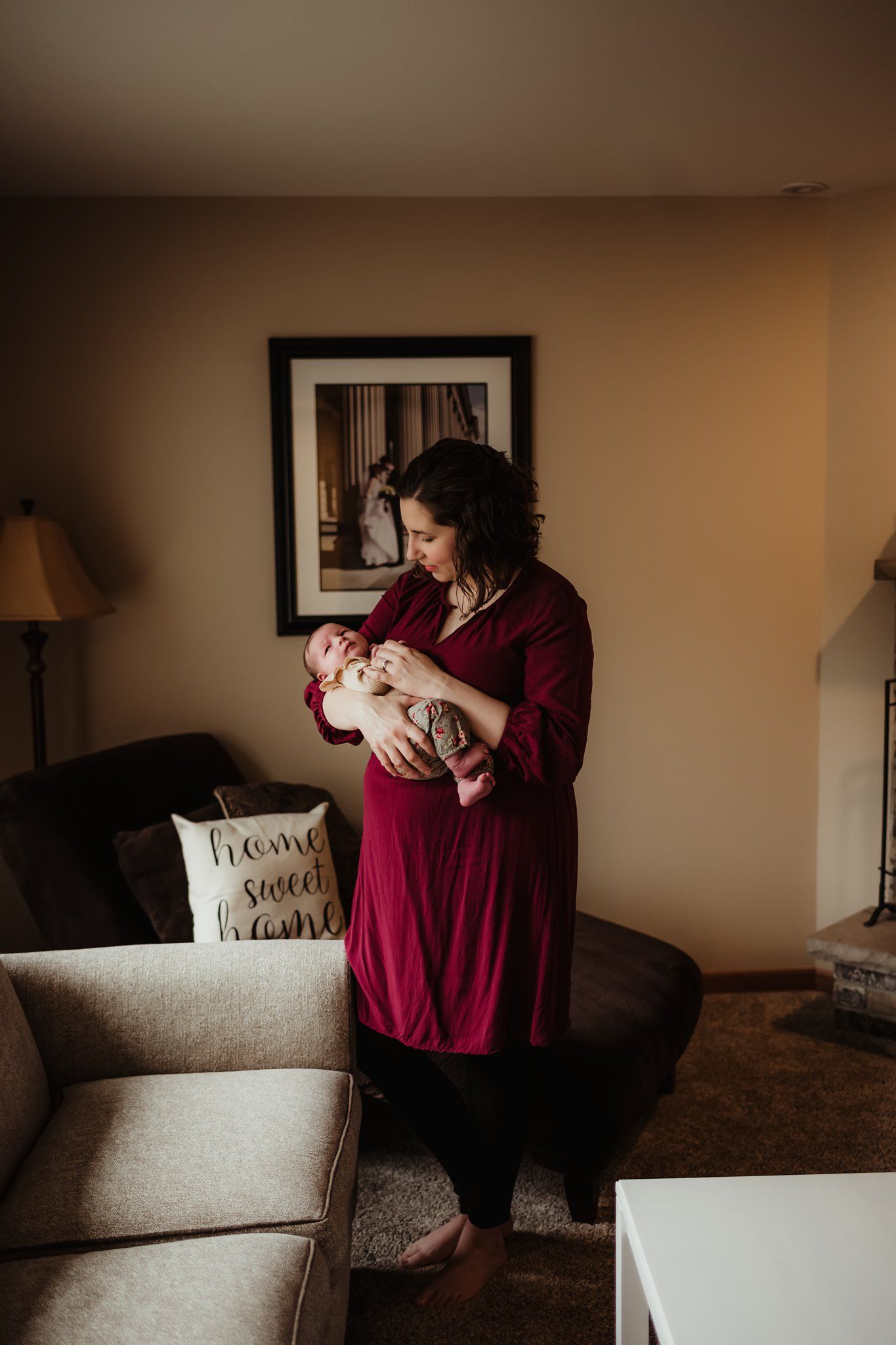 A person in a burgundy dress holds a newborn baby while standing in a warmly lit living room.