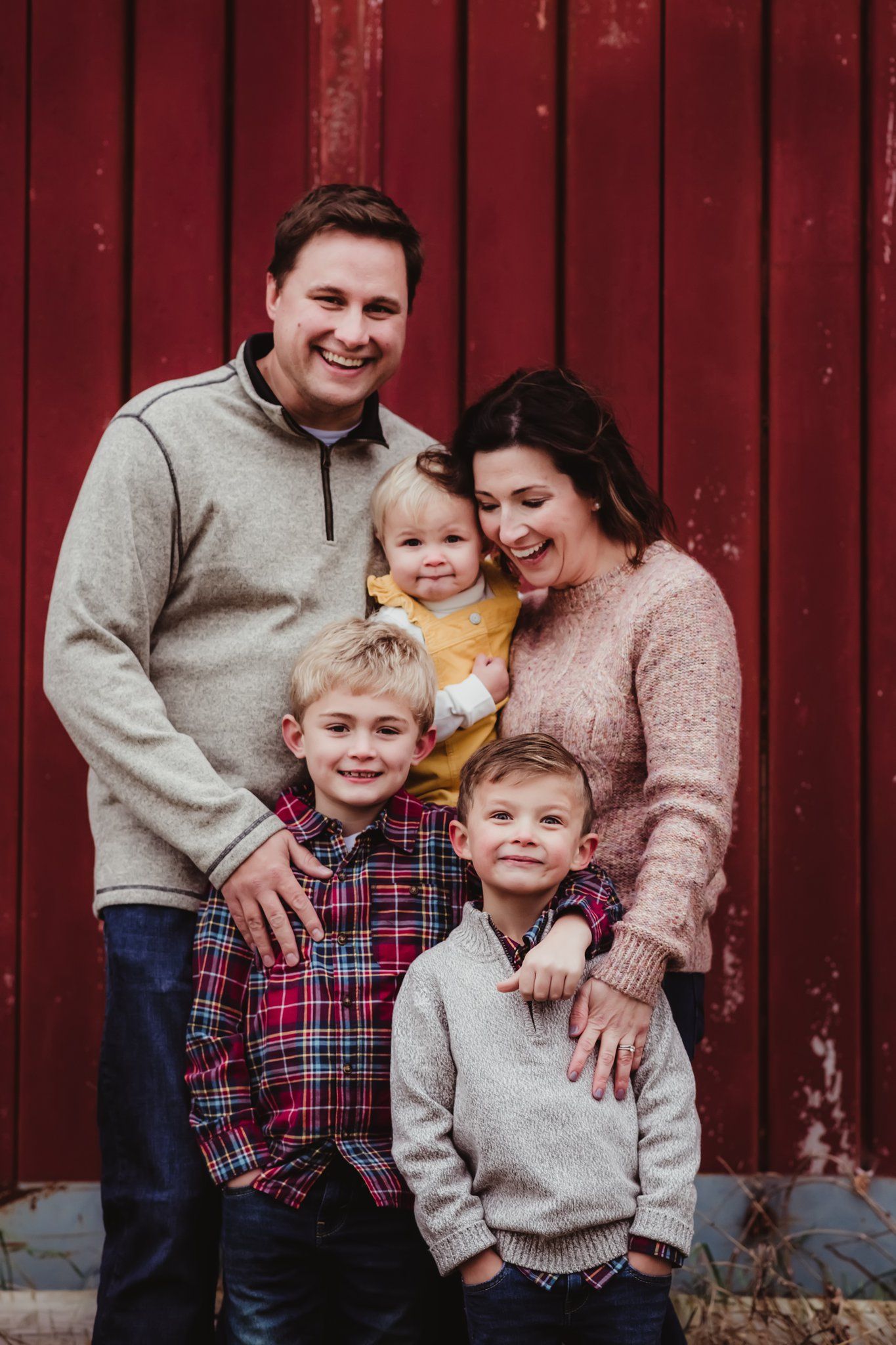 A cheerful family of five poses together against a red barn wall wearing coordinated casual outfits.