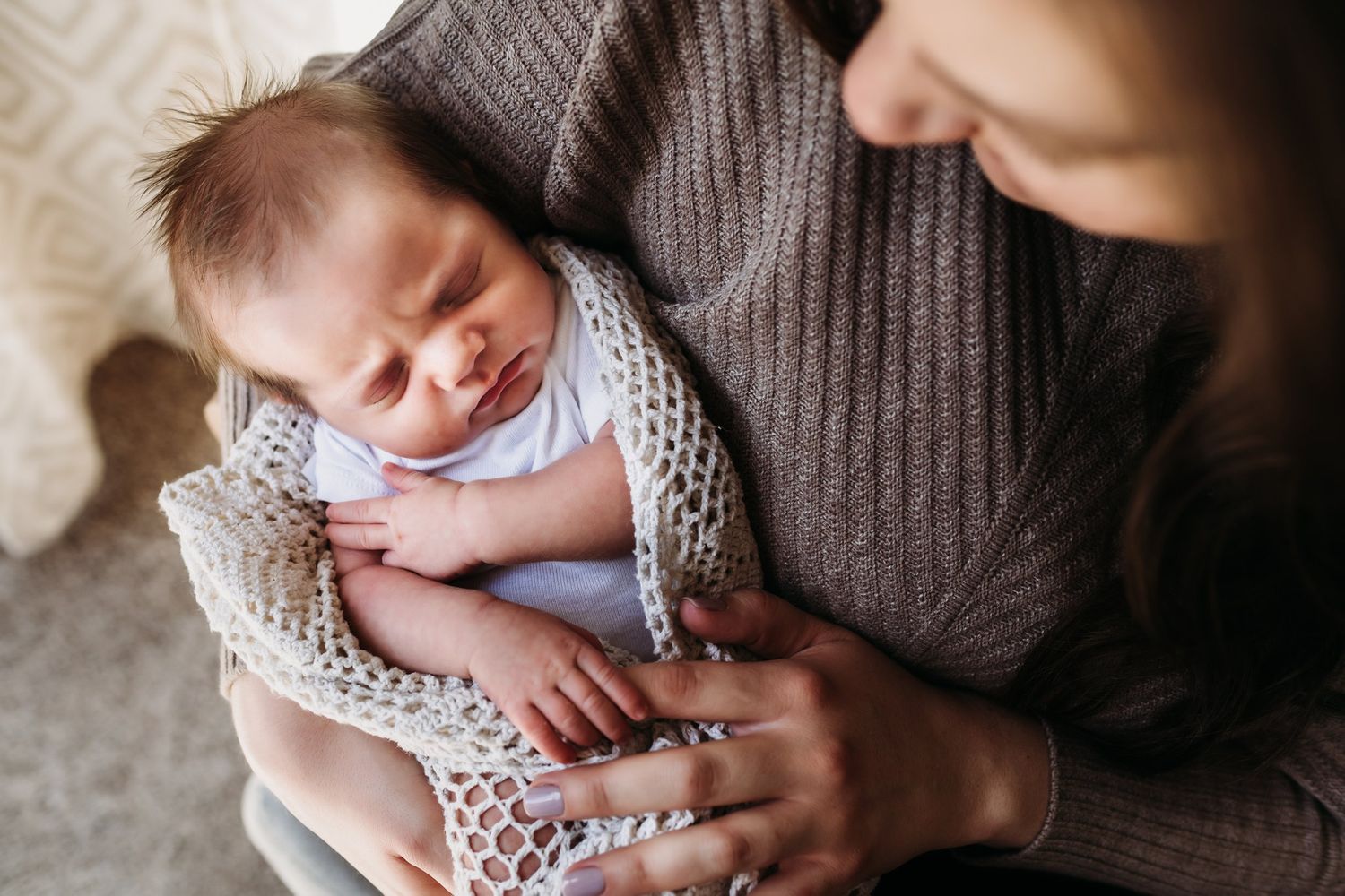 Close-up of sleeping newborn wrapped in cream knit blanket being held in brown sweater.