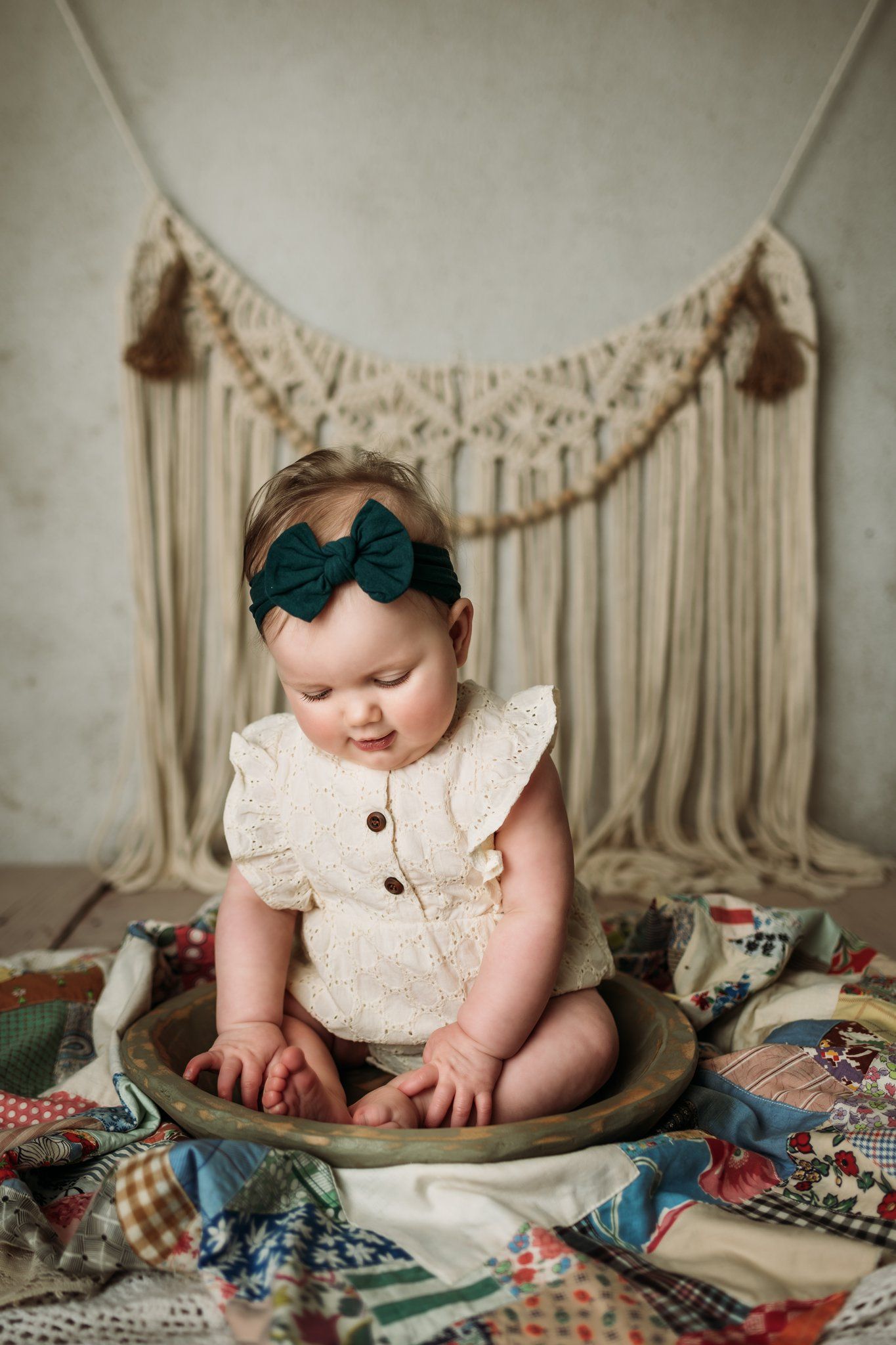 Baby wearing cream colored outfit with teal bow sits in wooden bowl on patchwork quilt.
