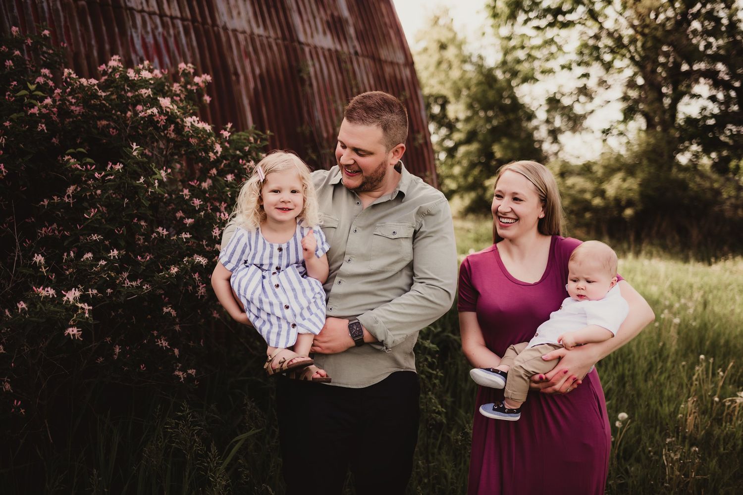 Natural light family portrait with rustic barn and flowering shrubs in background.