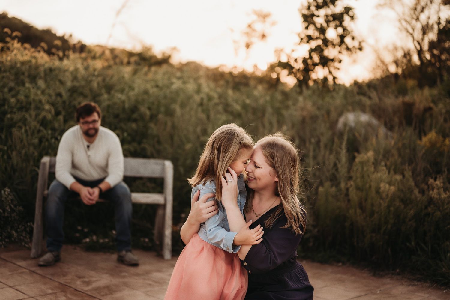 Three people share an intimate family moment in a sun-drenched field at golden hour.