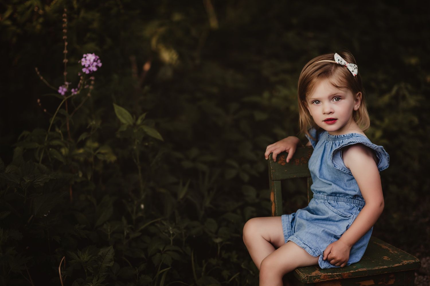 Candid shot of child in denim outfit sitting on wooden chair in garden setting.