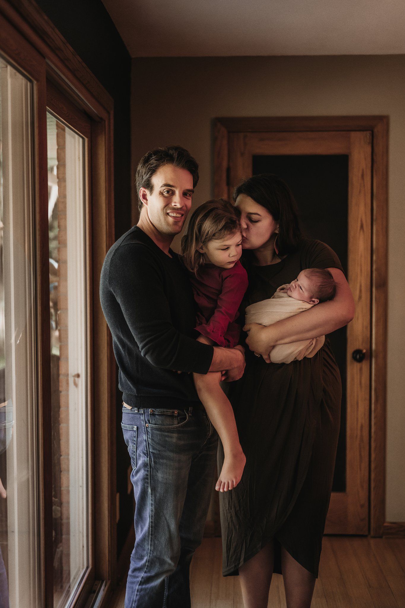Family of four poses by window with newborn baby and young child.