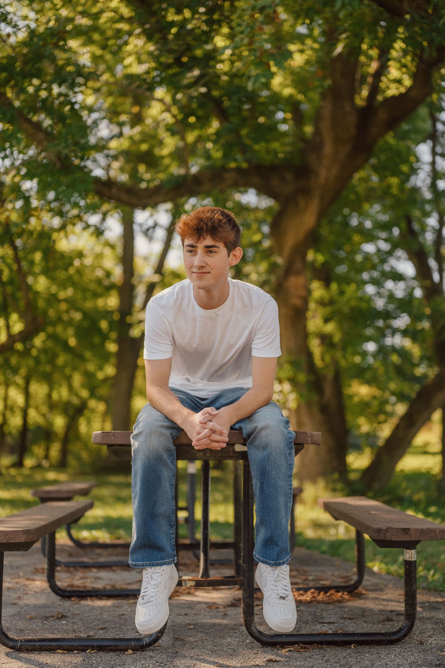 A person in a white t-shirt and jeans sits on a wooden picnic table in a sunny park setting.