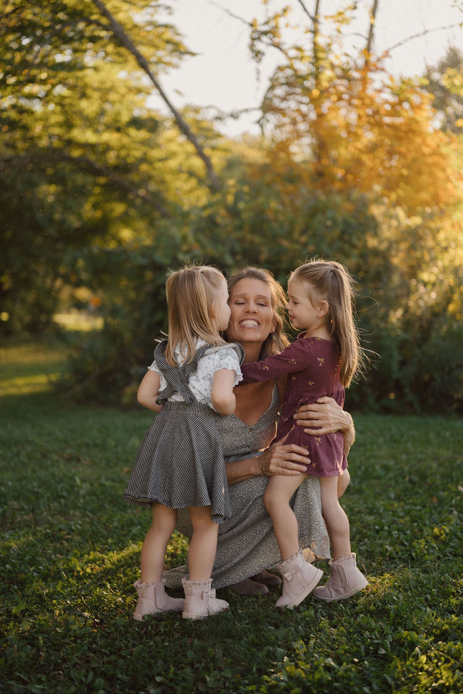 A tender family moment shared on grass during golden hour with autumn trees in the background.