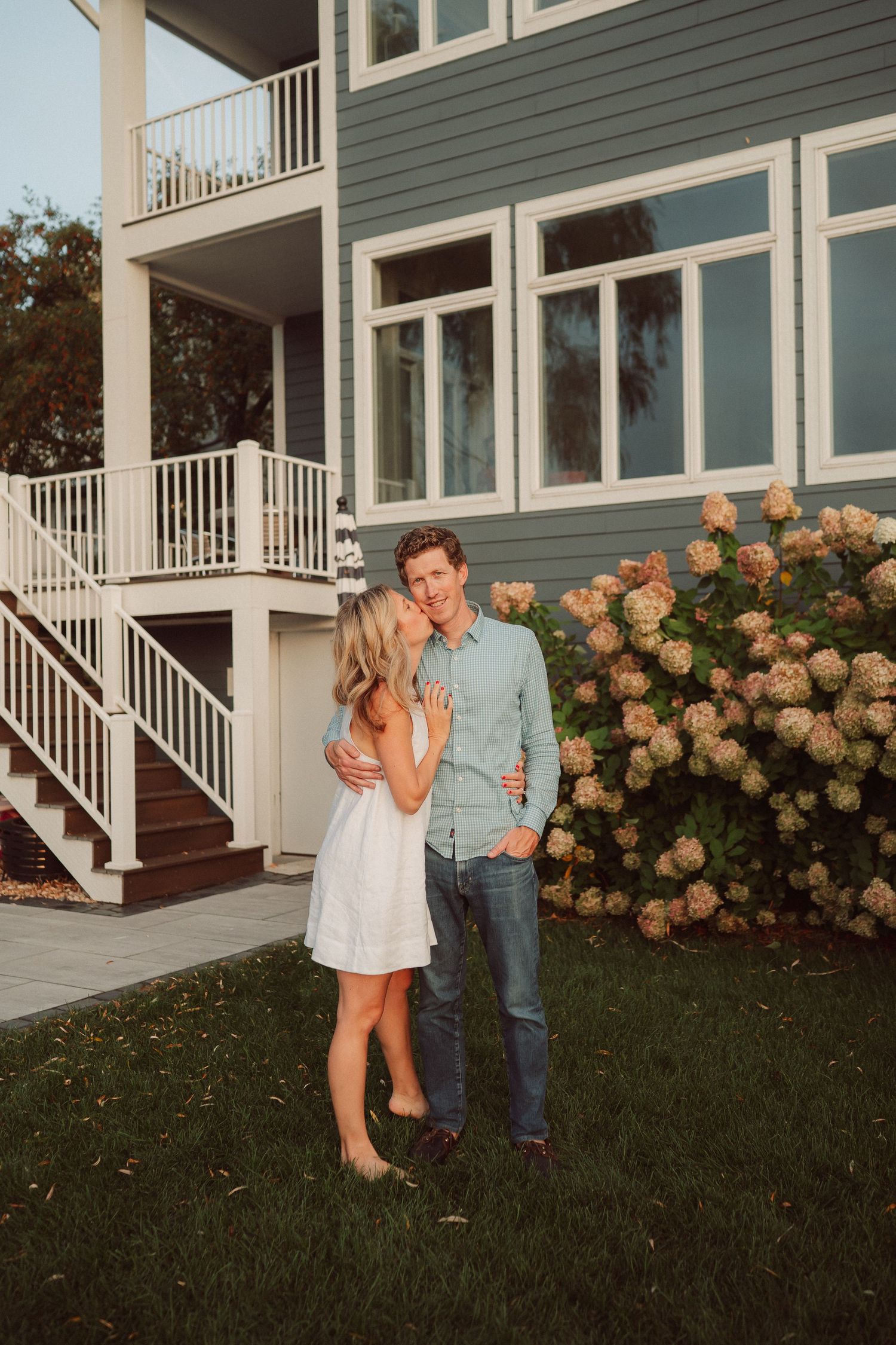 A couple dances playfully on the lawn outside a modern white and green house with blooming hydrangea bushes.