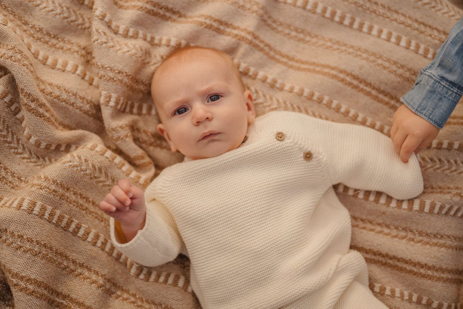 A baby lies on a patterned blanket while being gently wrapped in a cream-colored swaddle cloth.
