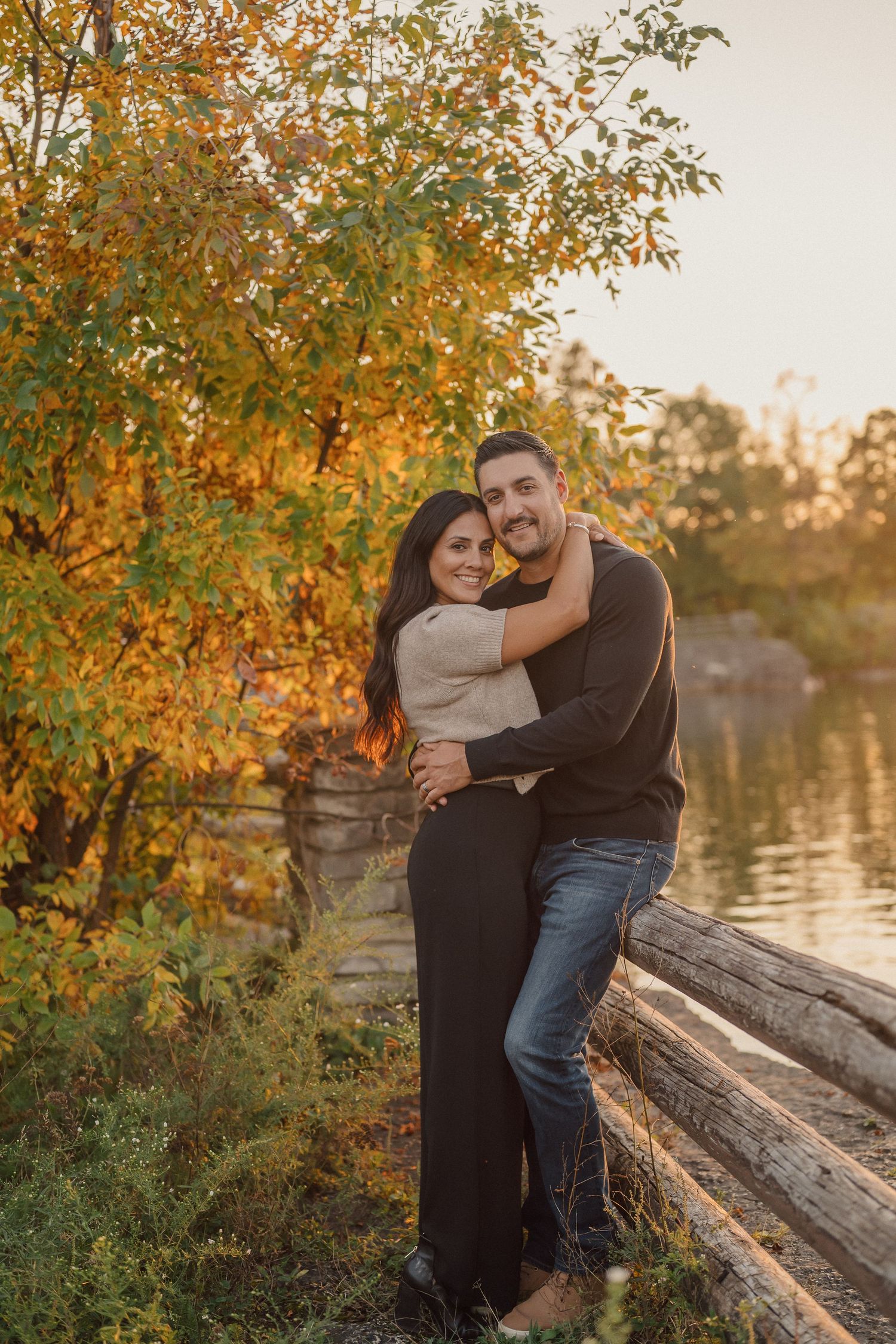 A couple embraces while sitting on a fallen log near a lake during autumn sunset with golden leaves overhead.
