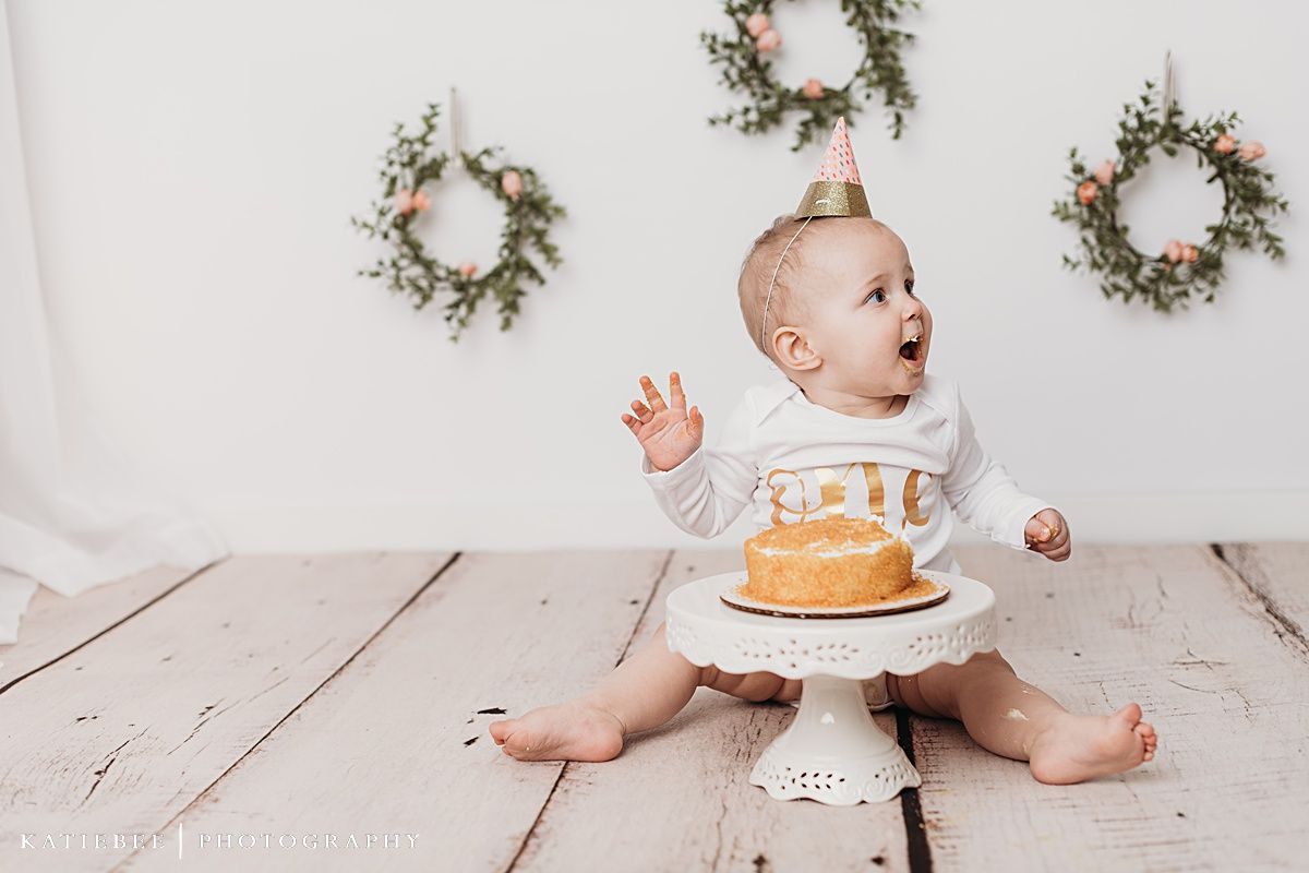 Baby wearing white onesie sitting near cake on pedestal with floral wreaths in background.