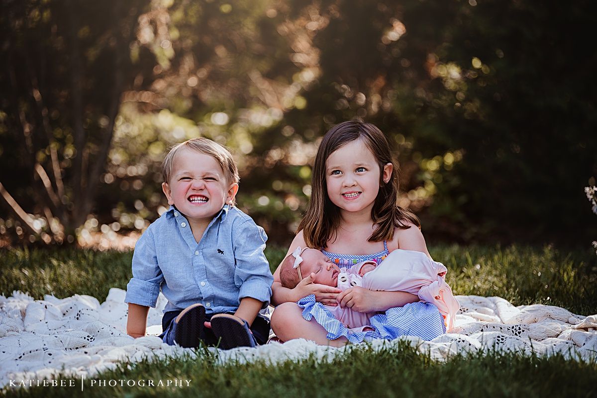 Siblings sitting together on white blanket outdoors during family photoshoot.