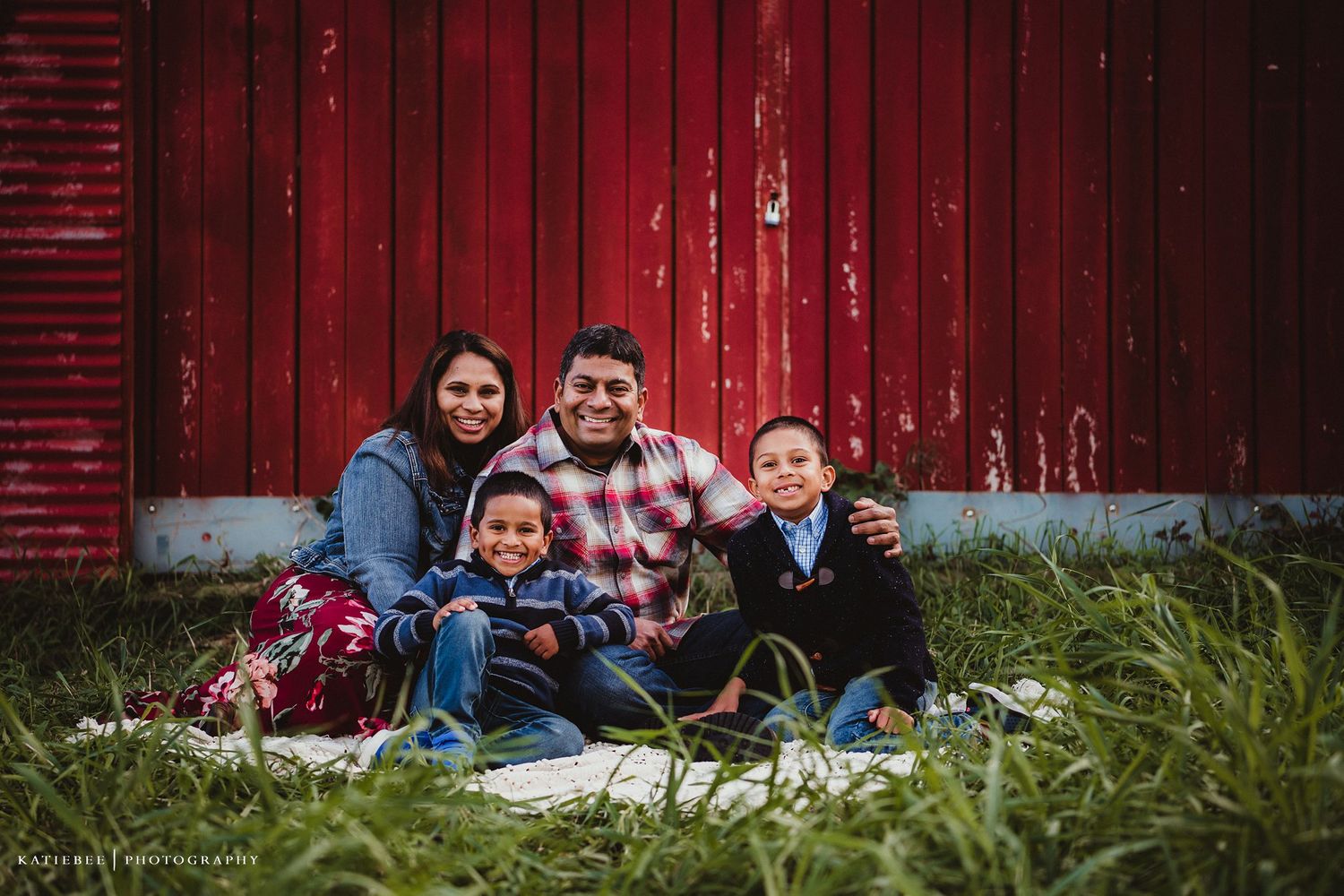 Family sitting together on grass against weathered red barn background.