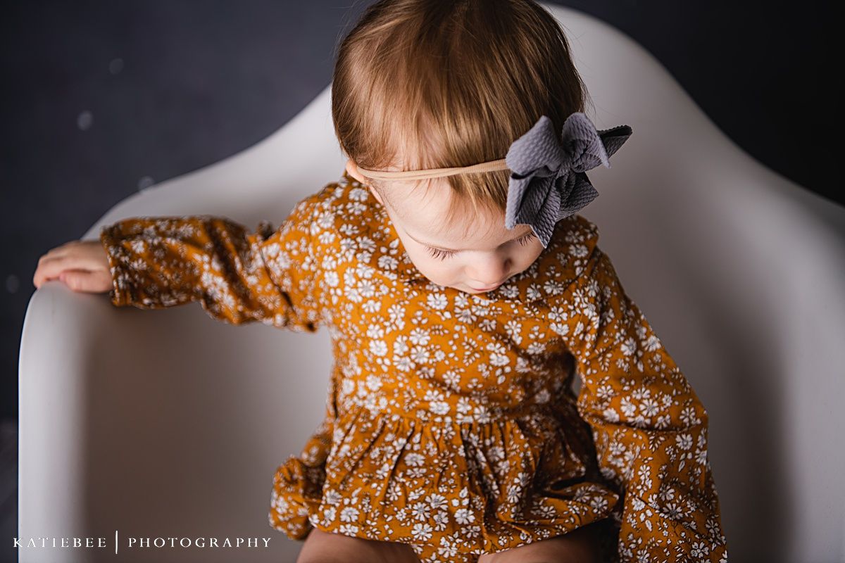 Close-up portrait of a toddler in a mustard floral dress and grey bow against dark background.