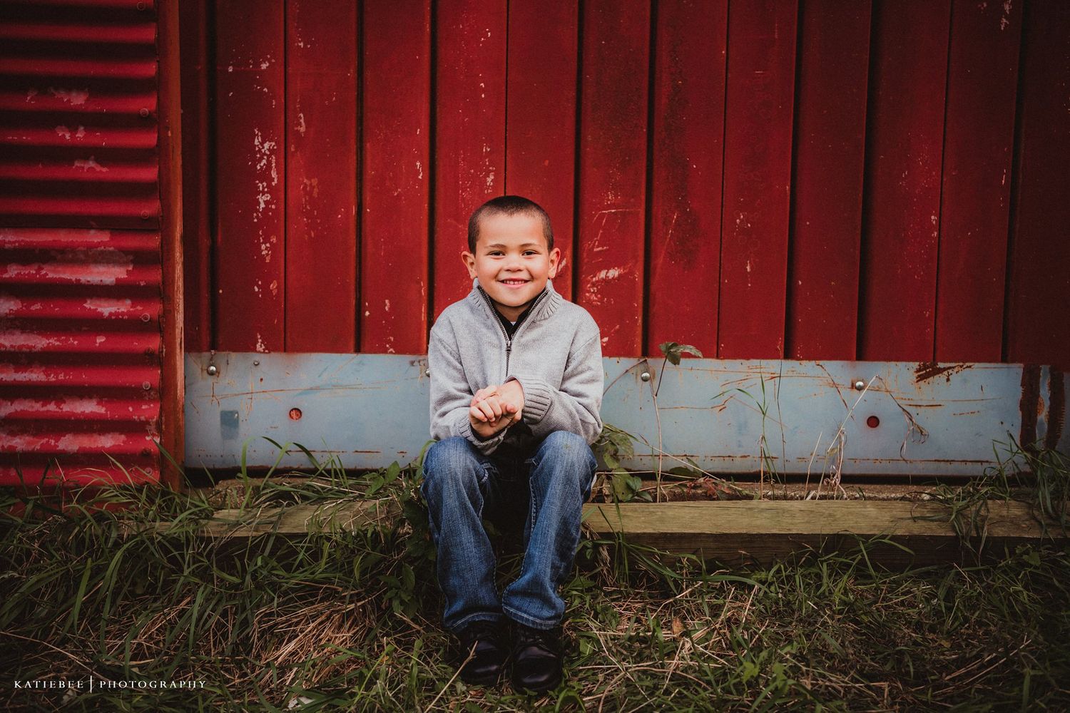 Child sitting casually against weathered red barn wall during outdoor photo session.