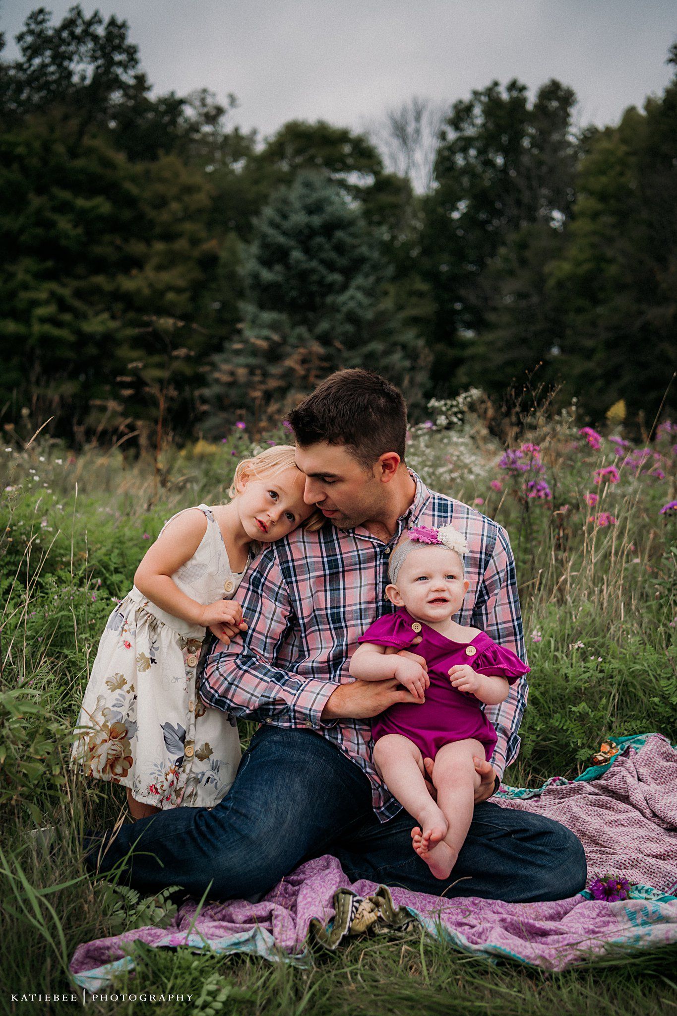 An intimate family portrait sitting on blanket in a meadow with purple wildflowers and evergreens.