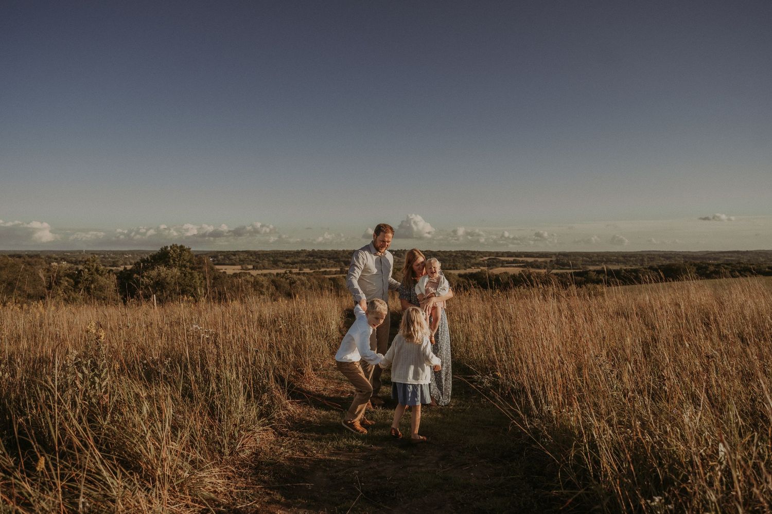 Family walking together on a dirt path through tall grass during sunset.