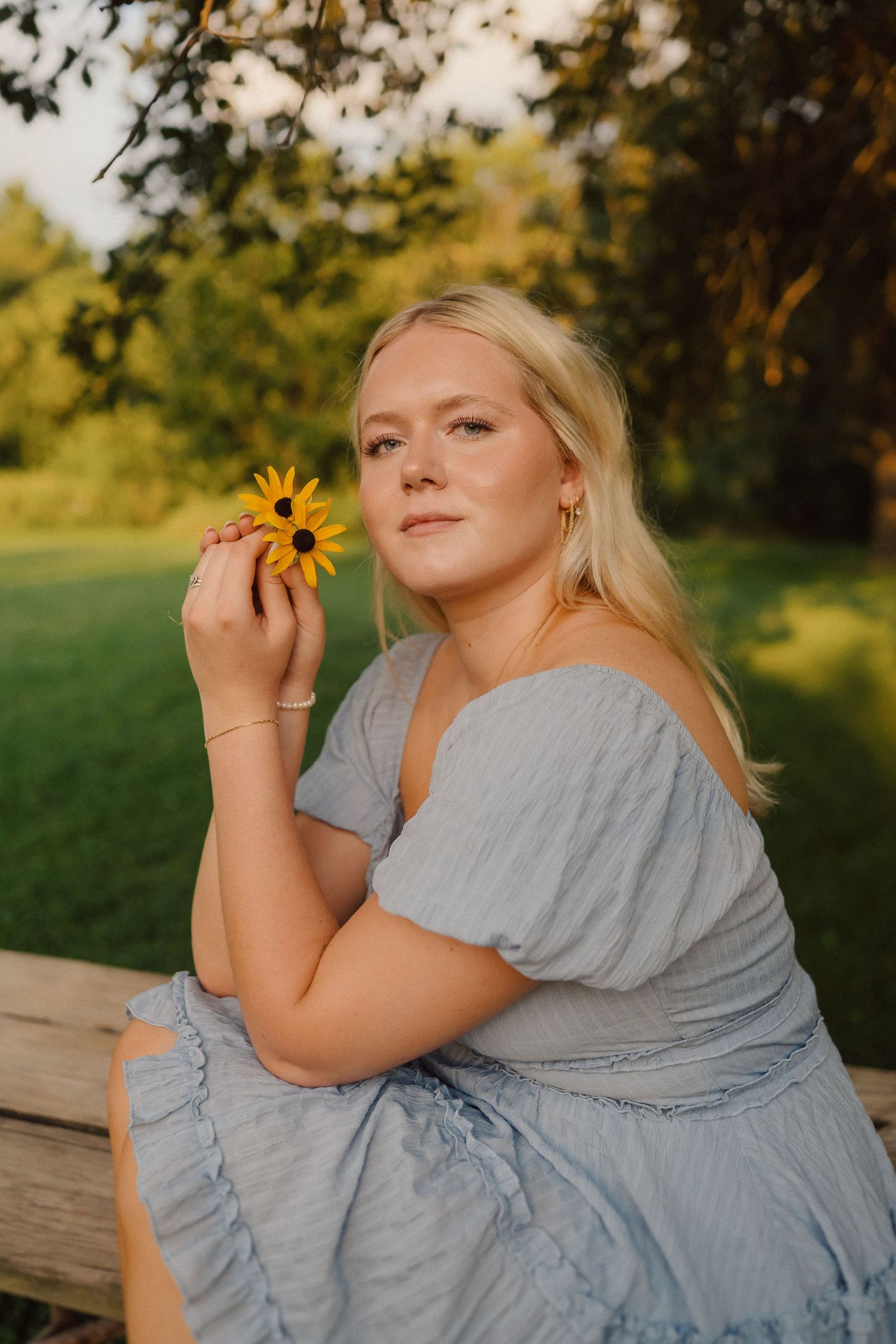 A relaxed summer portrait on a wooden bench with a person in a blue dress holding a sunflower at sunset.