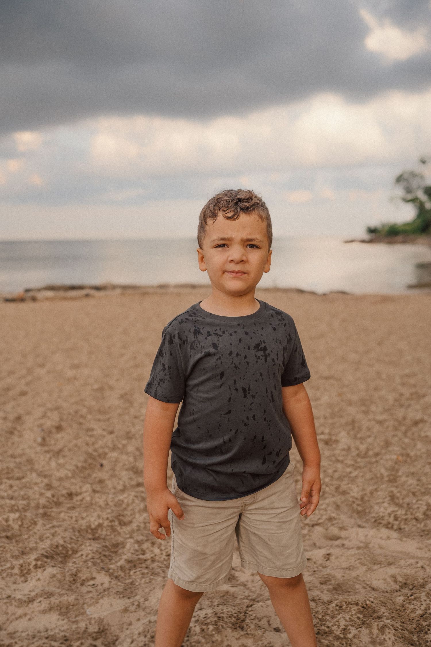 A child stands on a sandy beach with moody storm clouds overhead at dusk.