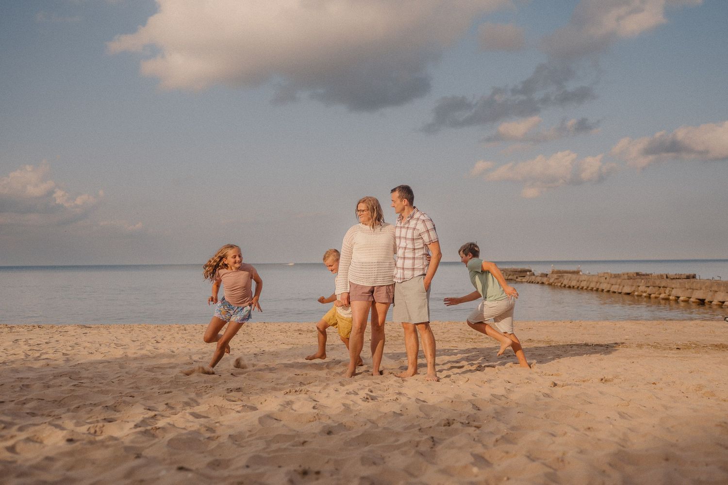 People running and playing together on a beach with a jetty and cloudy sky in the background.