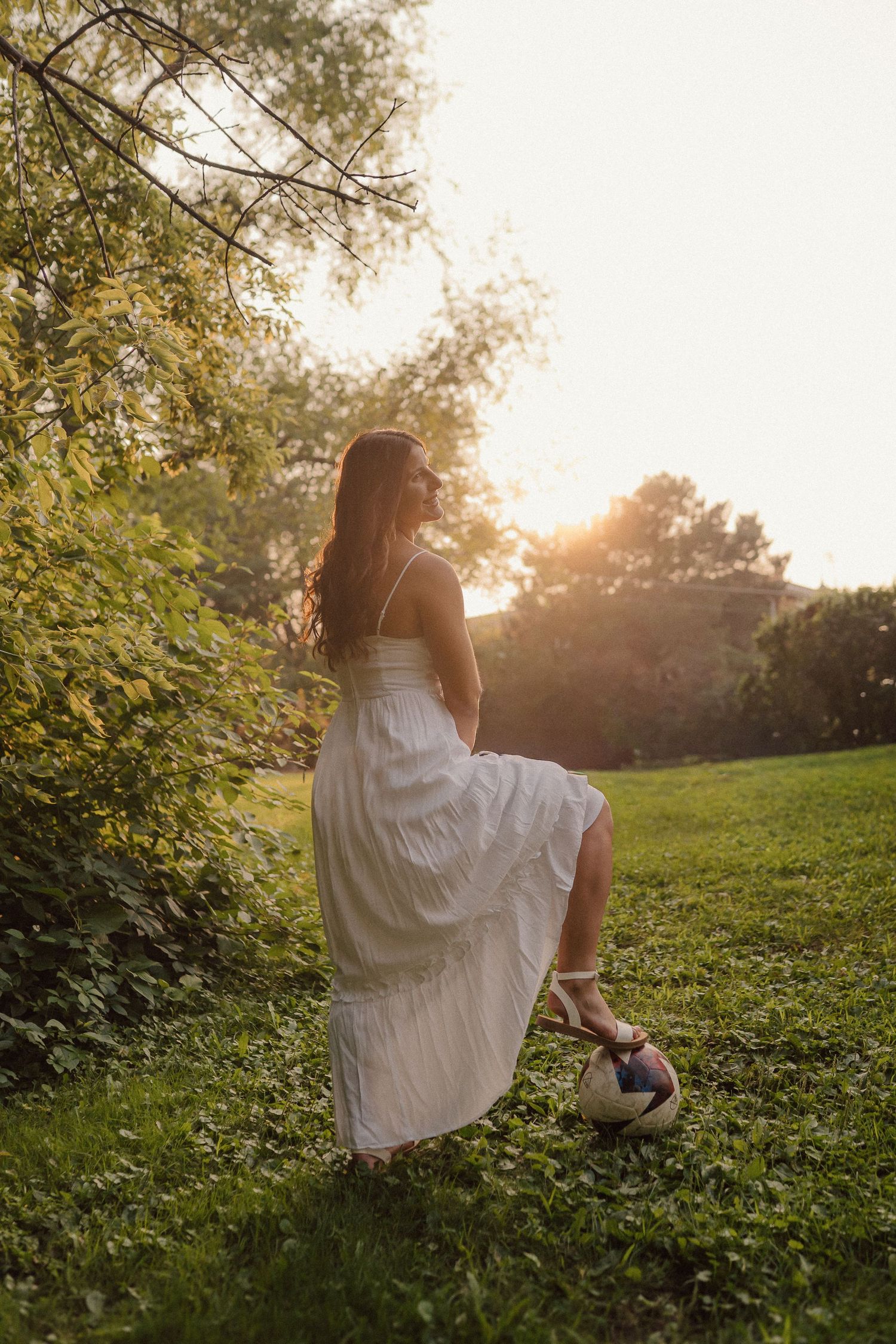 A person in a flowing white dress stands gracefully in a garden at golden hour with sunlight filtering through trees.