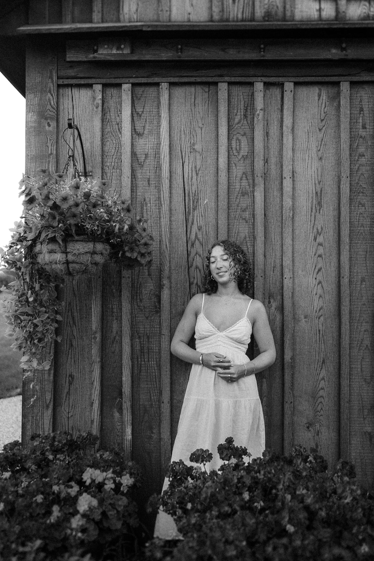 A person in a white summer dress poses against a rustic wooden fence with hanging plants and garden flowers below.