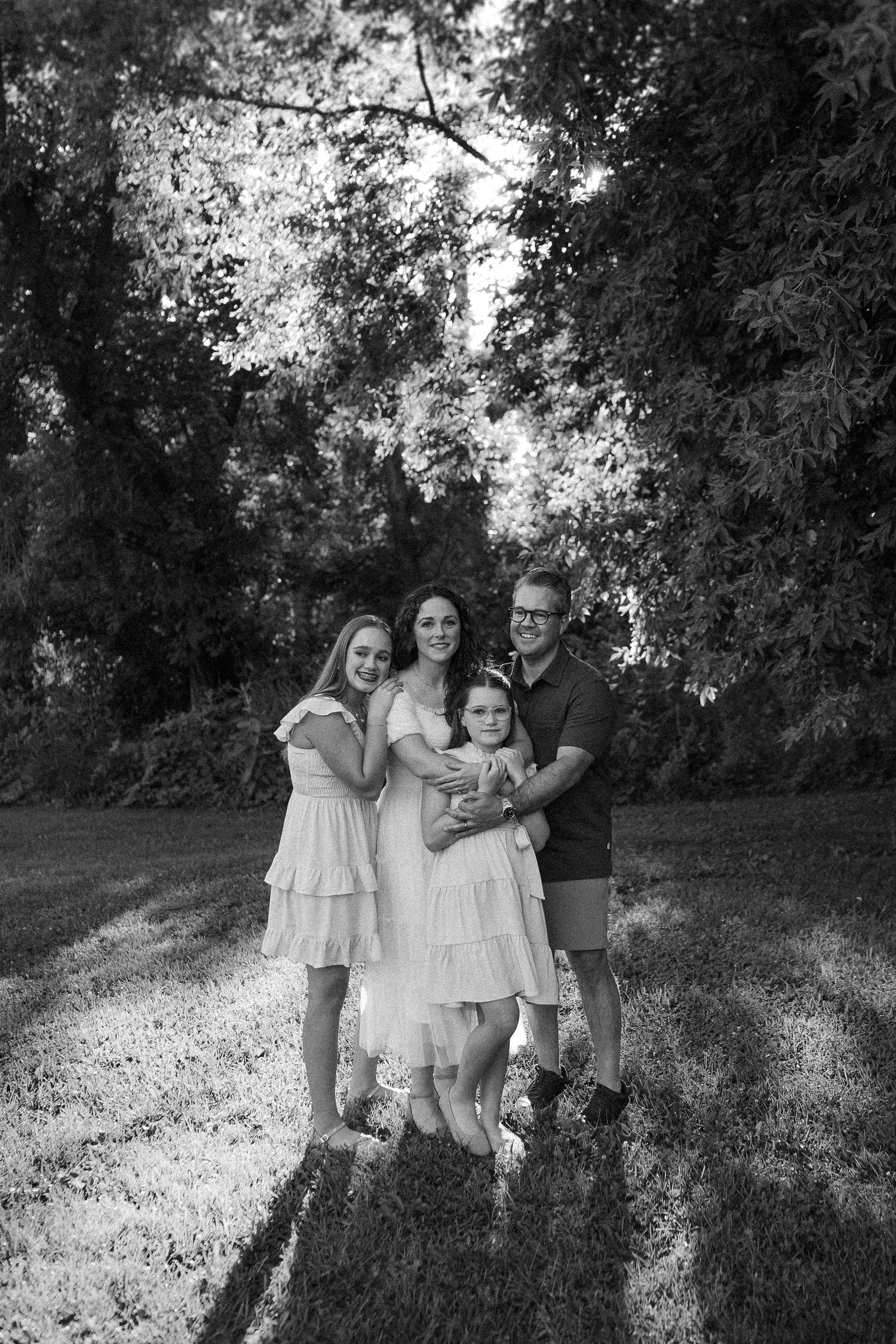 Black and white portrait of a family group embracing outdoors under trees.