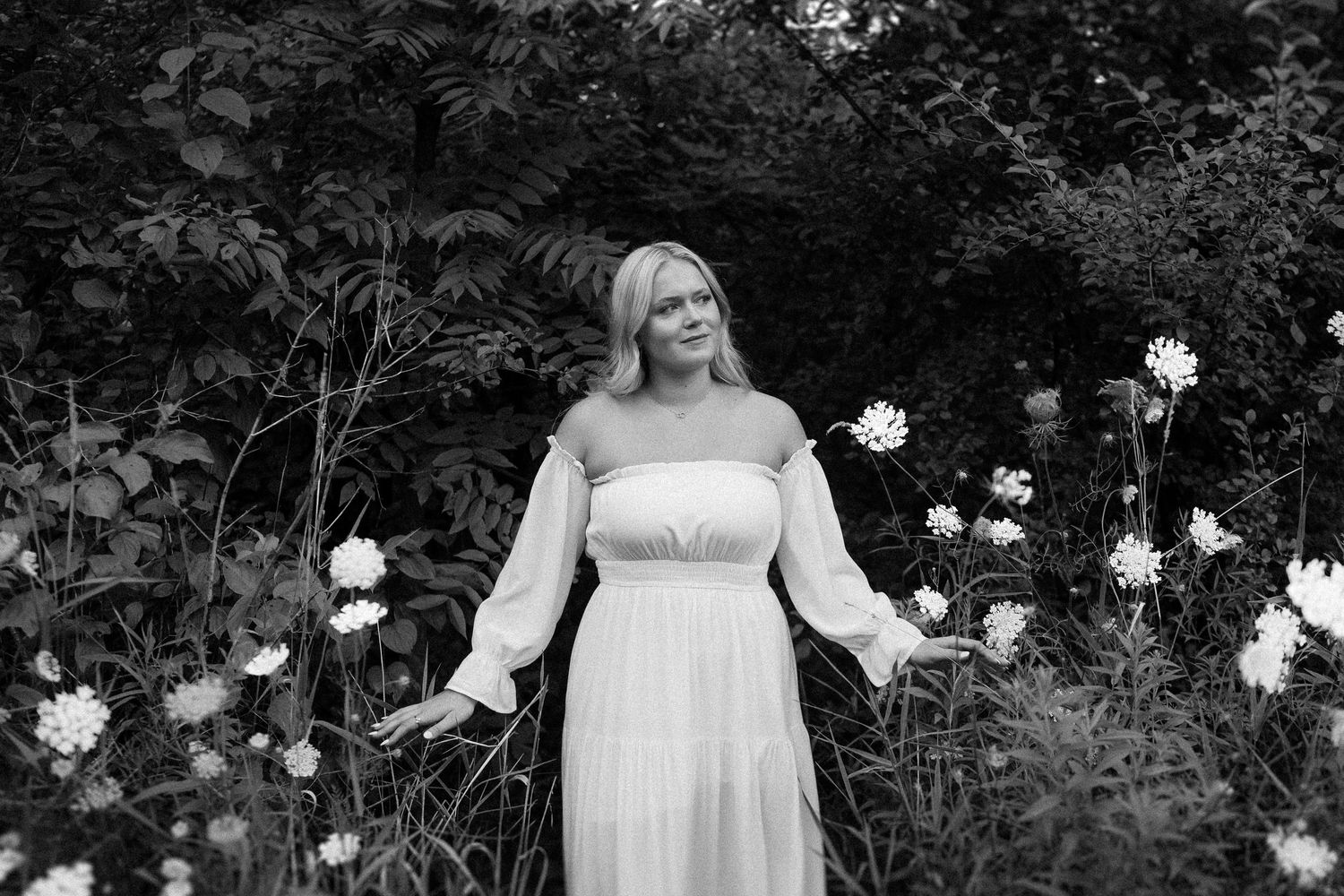 Person in white dress stands amid blooming white flowers and lush garden foliage in black and white.