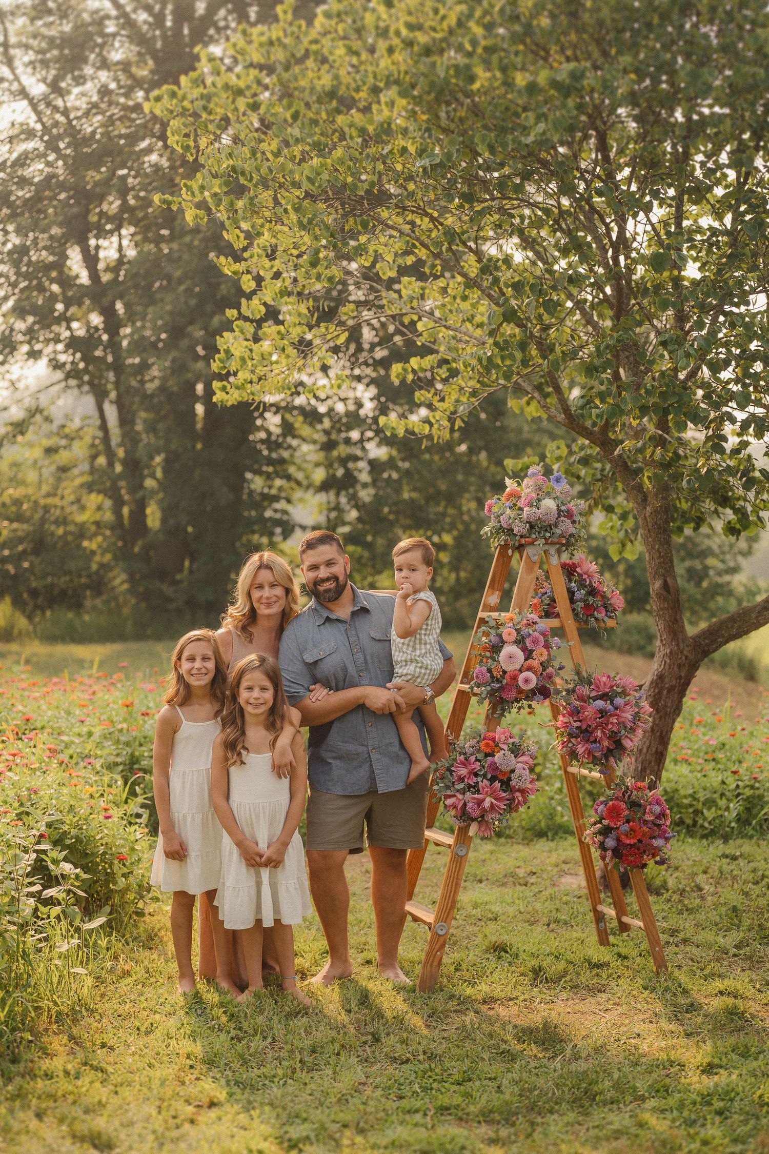 Family group portrait in casual attire near a wooden arch with pink floral decorations in garden setting.