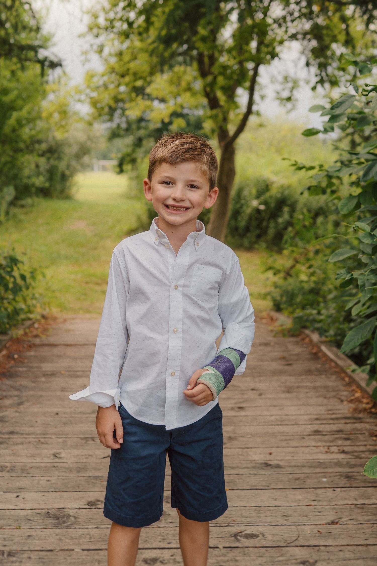 A young child stands smiling on a wooden pathway surrounded by green foliage on a sunny day.