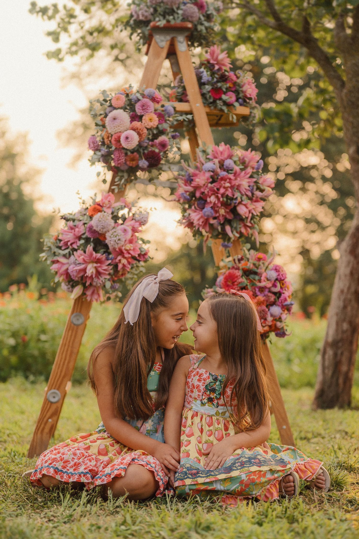 Two young friends in floral dresses sit together under a wooden arch decorated with pink flowers in a garden setting.