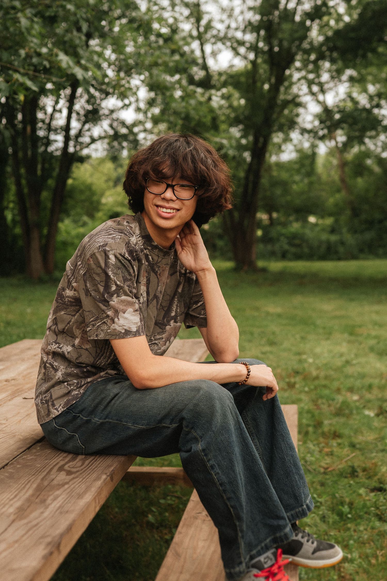 Young person in camo shirt and jeans sits casually on wooden bench in park-like outdoor setting.