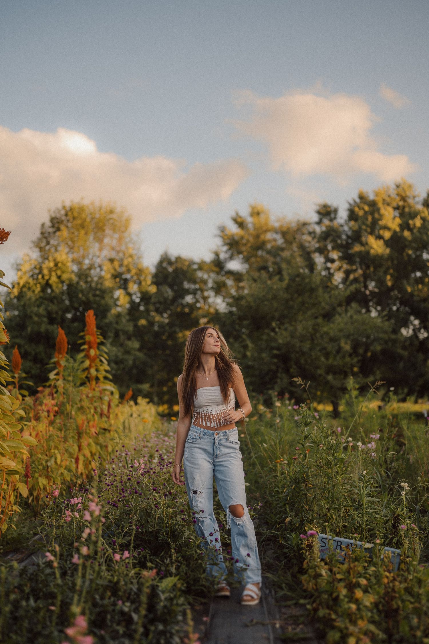 Young woman in ripped jeans standing among wildflowers at sunset.