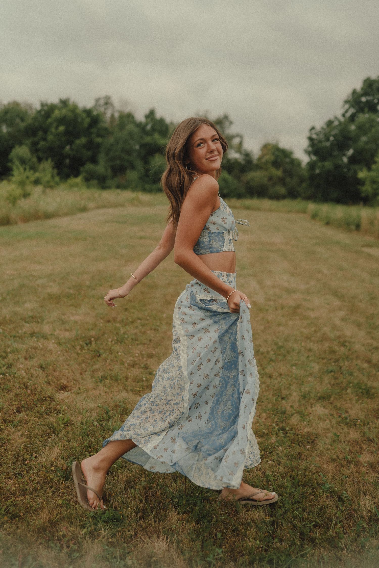 Smiling young woman in blue floral skirt set twirling in an open grassy field.