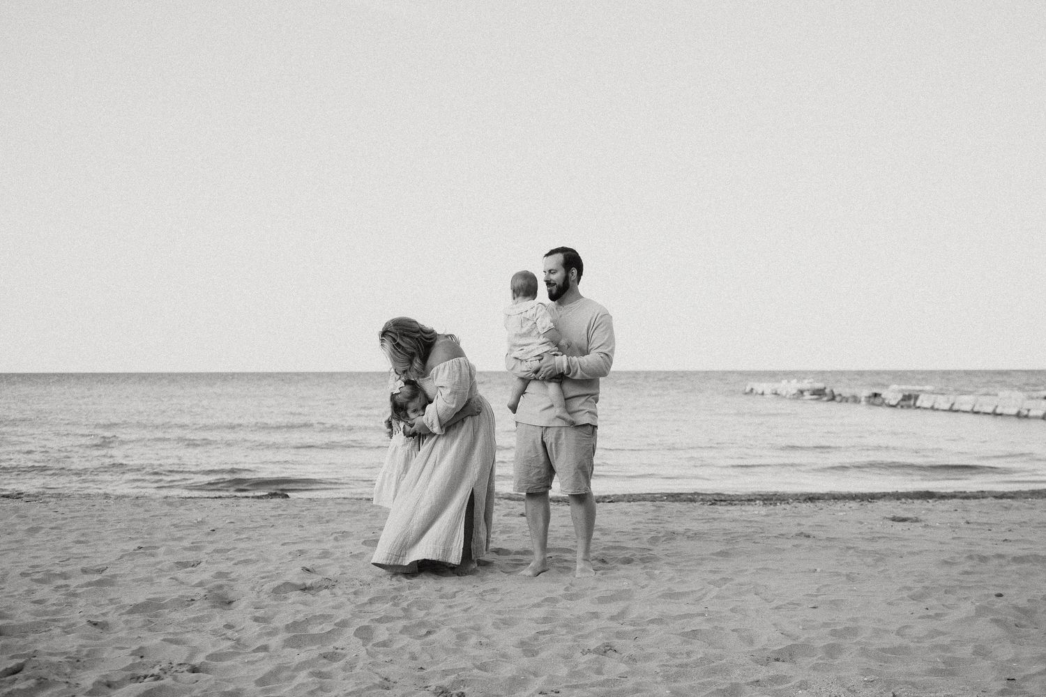 Black and white beach photo of family with two children, mother cradling baby while father stands nearby.
