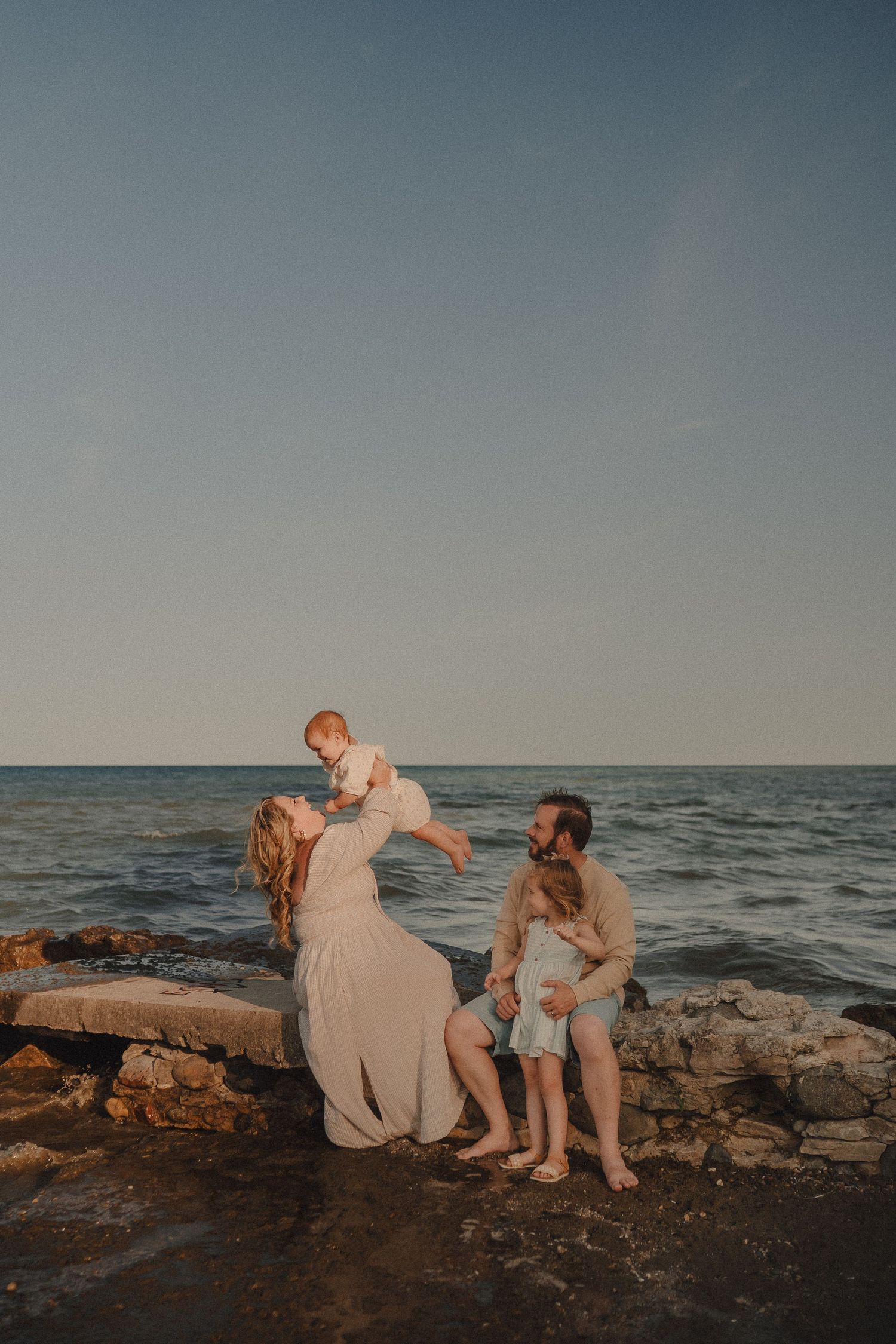 Family of four enjoying golden hour at rocky shoreline, mother lifting baby while father sits with toddler.