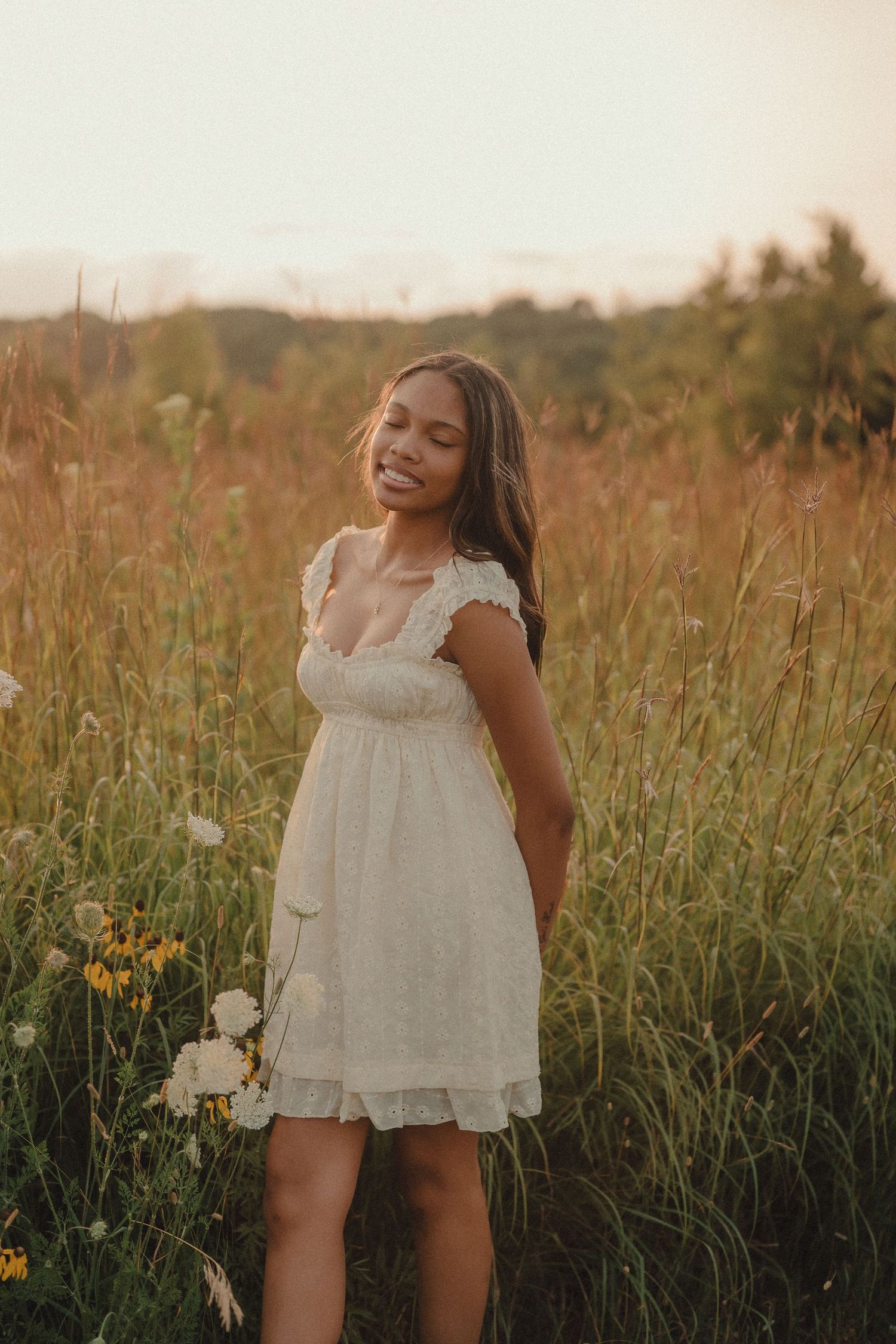 Young woman in white lace dress holding wildflowers, smiling in a sunlit meadow at golden hour.