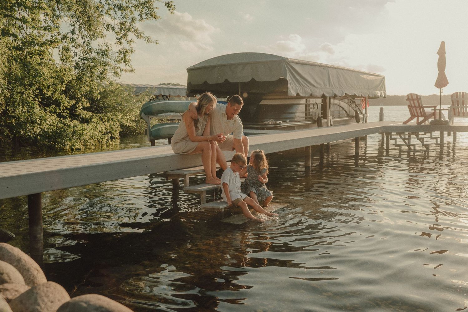 Family sitting on a dock with feet in the water, enjoying a sunny day by the lake near a waterfront restaurant.