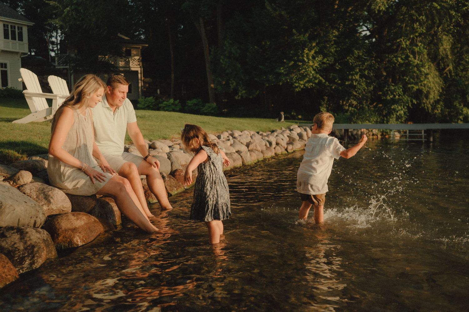 Family sitting on rocky shore at golden hour while children splash in calm lake water.