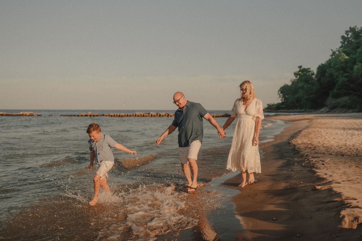 A family of three laughs and splashes along a sunlit beach shoreline, holding hands near the water.