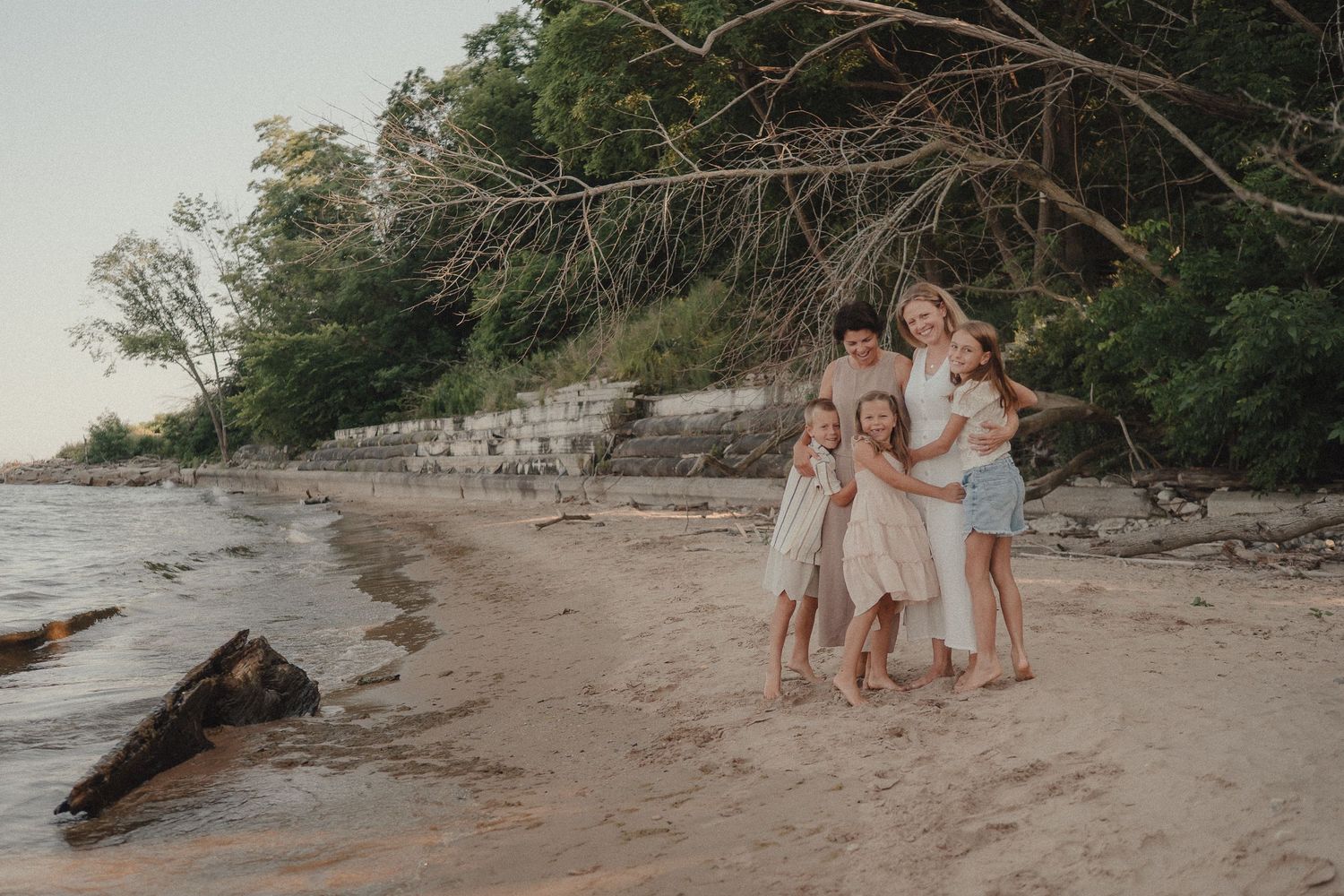 Happy family group hug on sandy beach with rocky cliff and fallen trees in background.