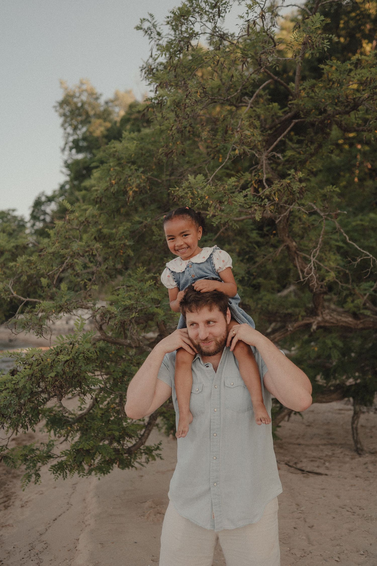 A man carries a smiling young girl on his shoulders outdoors near lush green trees on a sandy path.