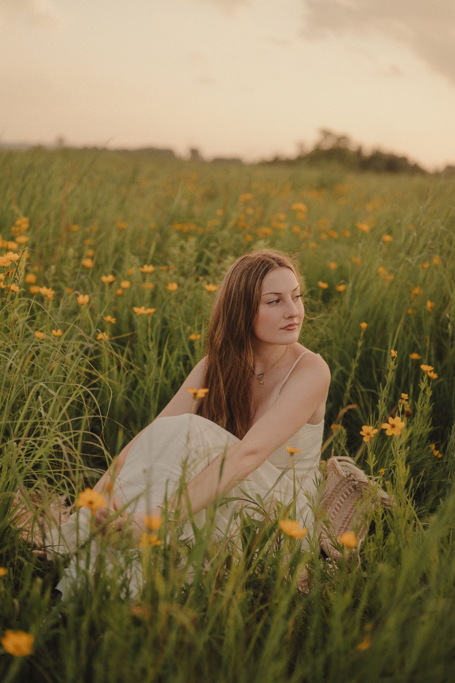 Young woman in a white dress sits among tall green grass and yellow wildflowers at sunset.