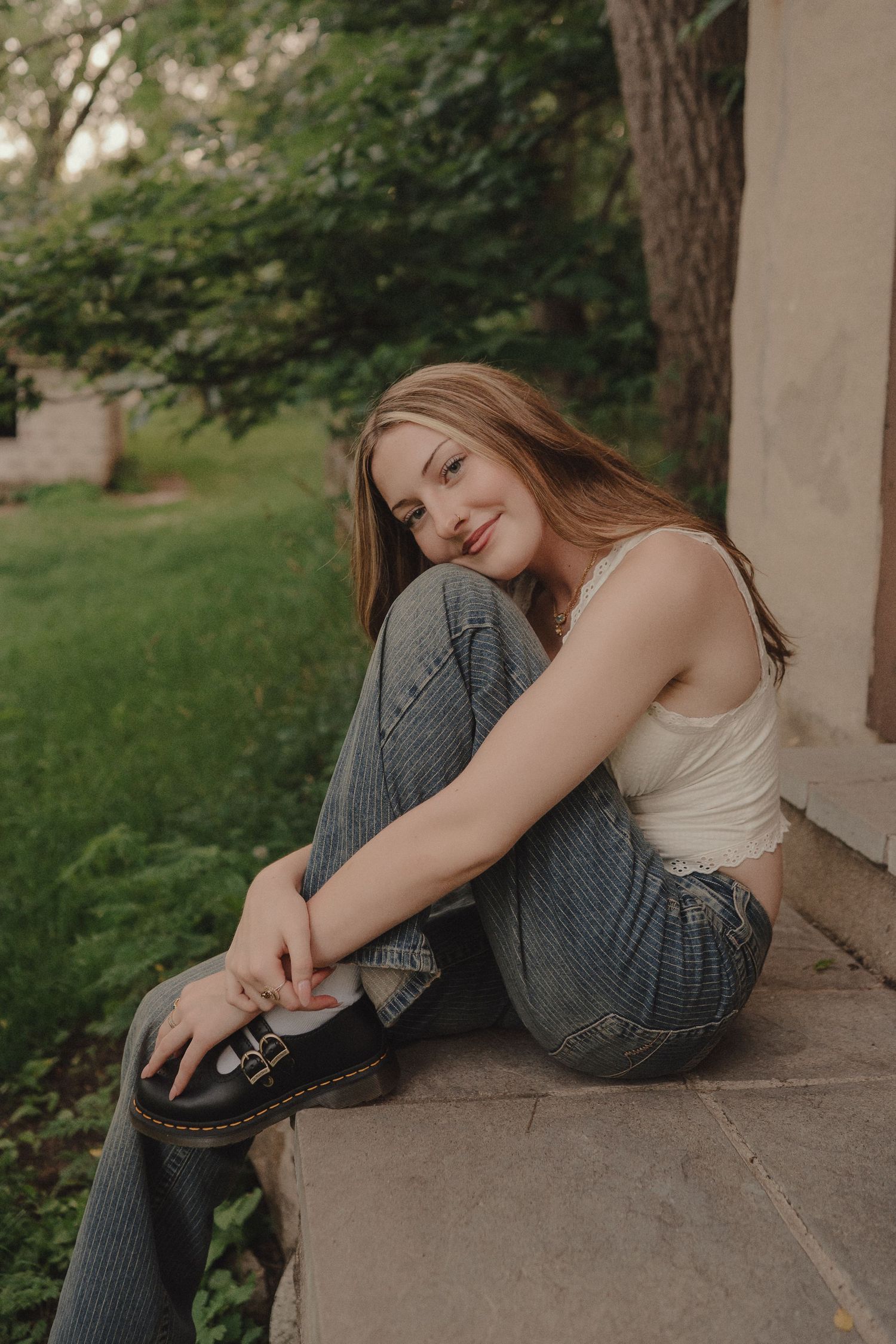 Young woman in jeans and white tank top sitting on stone steps outdoors, smiling softly with greenery background.