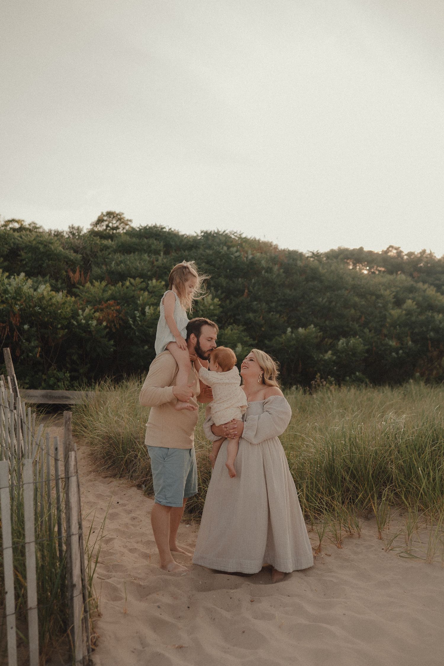 Happy family of four on sandy beach path, father with child on shoulders, mother holding baby.