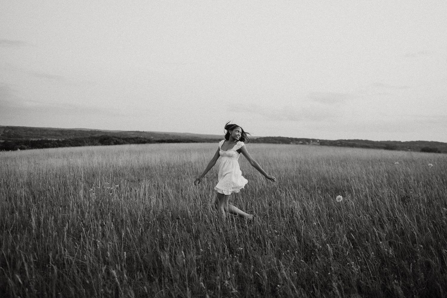 Black and white photo of a girl in white dress joyfully running through a vast open field of tall grass.