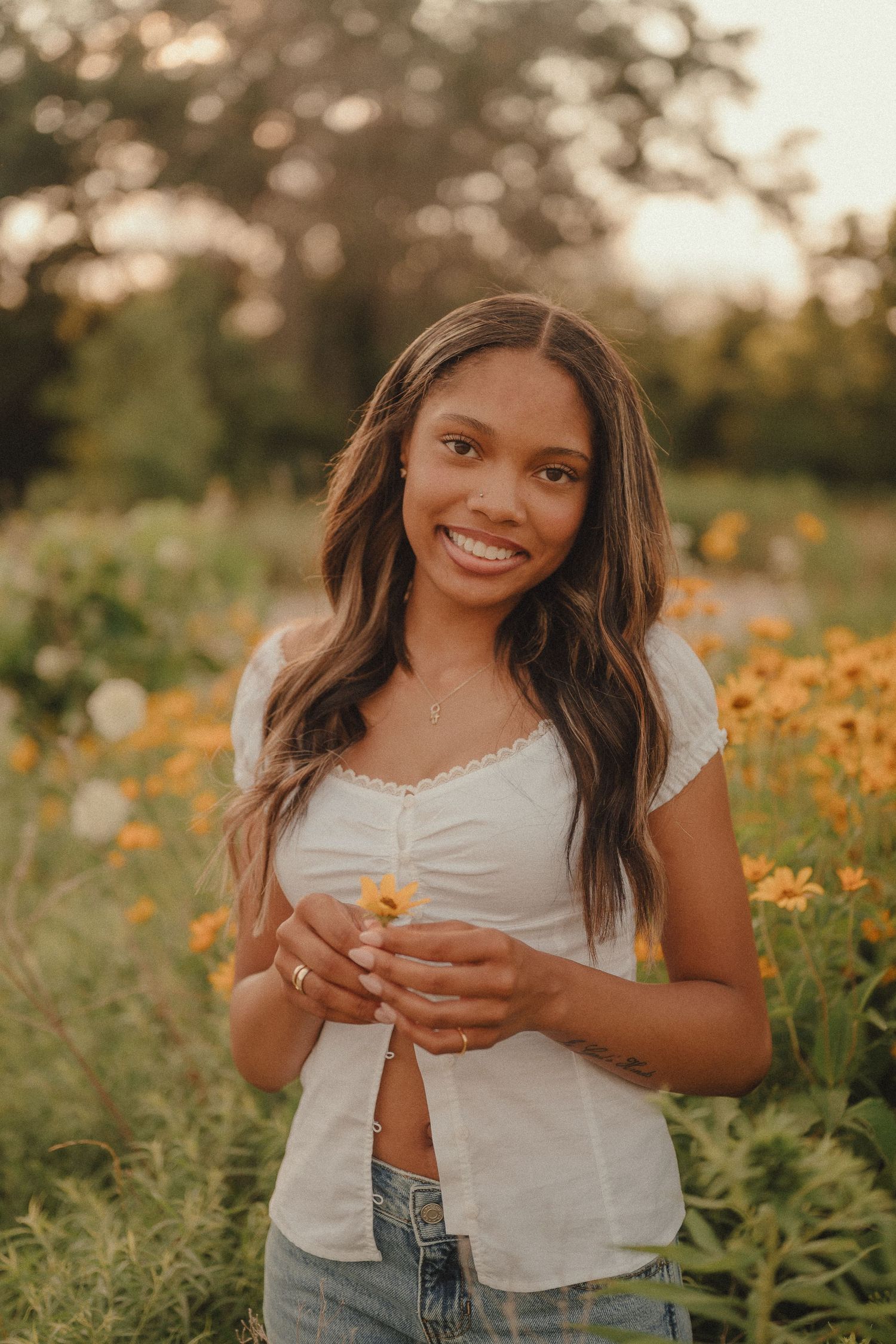 Smiling young woman holding yellow wildflowers in a sunny field, wearing white top and jeans.