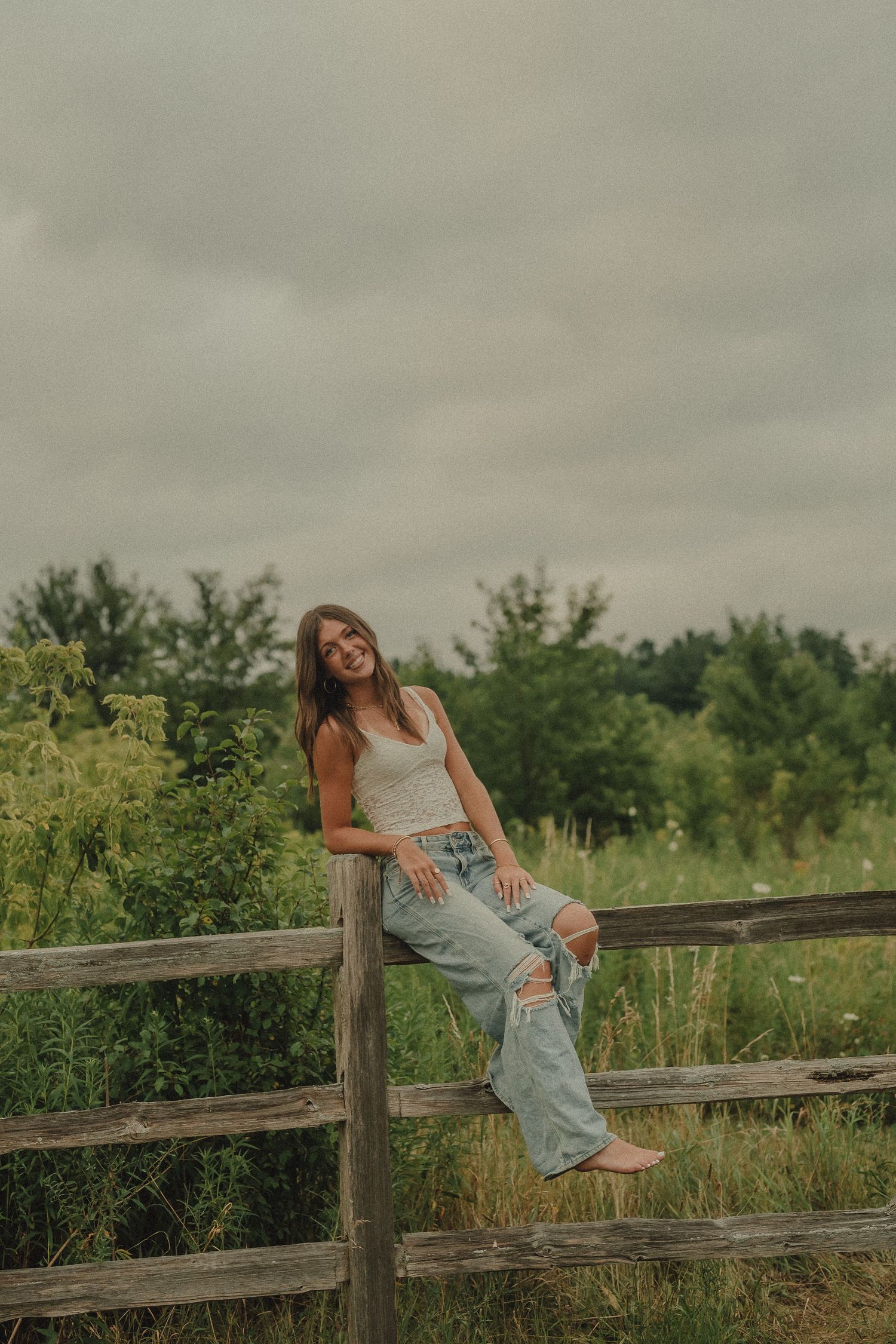 Young woman in white tank top and ripped jeans sitting on a rustic wooden fence in a green field.